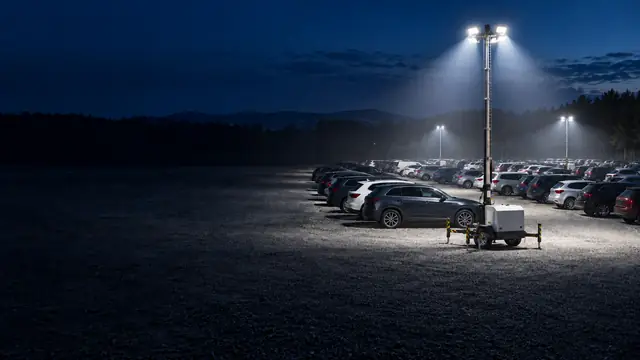 Mobiler LED-Lichtmast auf Anhänger leuchtet einen Schotterparkplatz mit Reihen parkender Autos bei Nacht aus